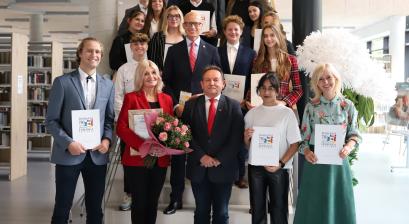 Grupa osób: Członkowie Młodzieżowej Rady Miasta Lublińca, Burmistrz, Zastępca Burmistrza, Przewodniczący Rady Miejskiej, Opiekun Młodzieżowej rady i zaproszeni goście, stojąca na schodach w Miejsko-Powiatowej Bibliotece Publicznej w Lublińcu
