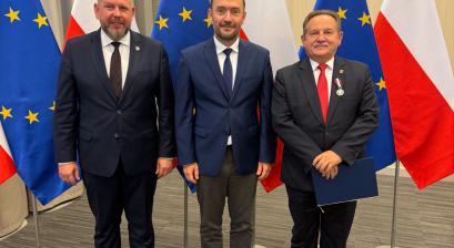 Burmistrz oraz osoby wręczające na tle flag 