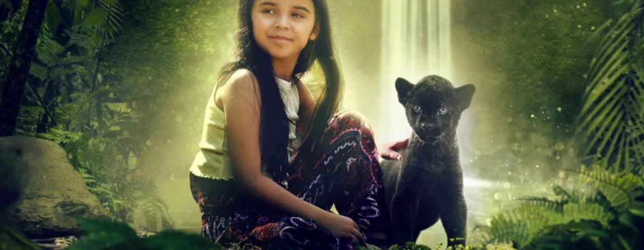 Dziewczynka siedząca w dżungli na tle wodospadu. U jej prawego boku siedzi mały czarny Jaguar