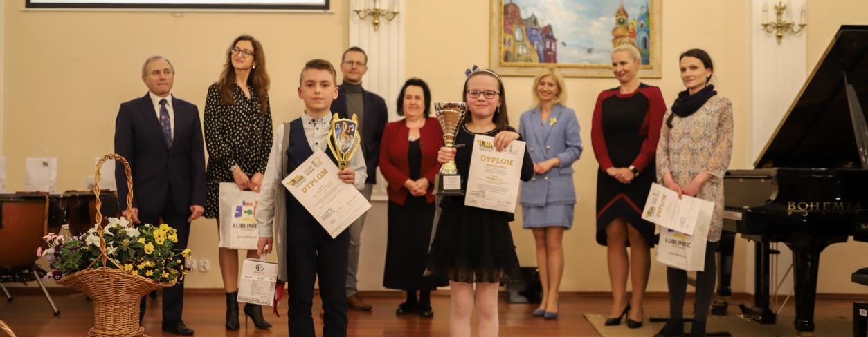 sala koncertowa szkoły muzycznej w Lublińcu na scenie stwoji dwójka dzieci zwycięsców Grand Prix Festiwalu dla Pianistów. Dzieci trzymają w rękach dyplomy i puchary, za nimi stoją członkowie komisjii konkursowej oraz wiceburmistrz Anna Jonczyk-Drzymała Lublińca wraz z przedstawicielem Rady Powiaty Panią Naczyńską