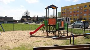 plac zabaw na skrzyżowaniu ul Piasecznej i Opolskiej na pierwszym palnie widać wieżyczkę drewnianego domku zabaw z zjeżdżalnią i przyłączonymi huśtawkami z widokiem bloków osiedla 3 maja w tle